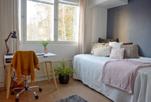 Särkiniementie 18 asunnon makuuhuone, jossa on vaalealla päiväpeitolla ja tyynyillä peitelty sänky sekä ikkunan edessä valkoinen työpöytä ja huonekasveja. The bedroom in Särkiniementie 18 has a bed covered with a light bedspread and pillows, a white desk and houseplants in front of the window.